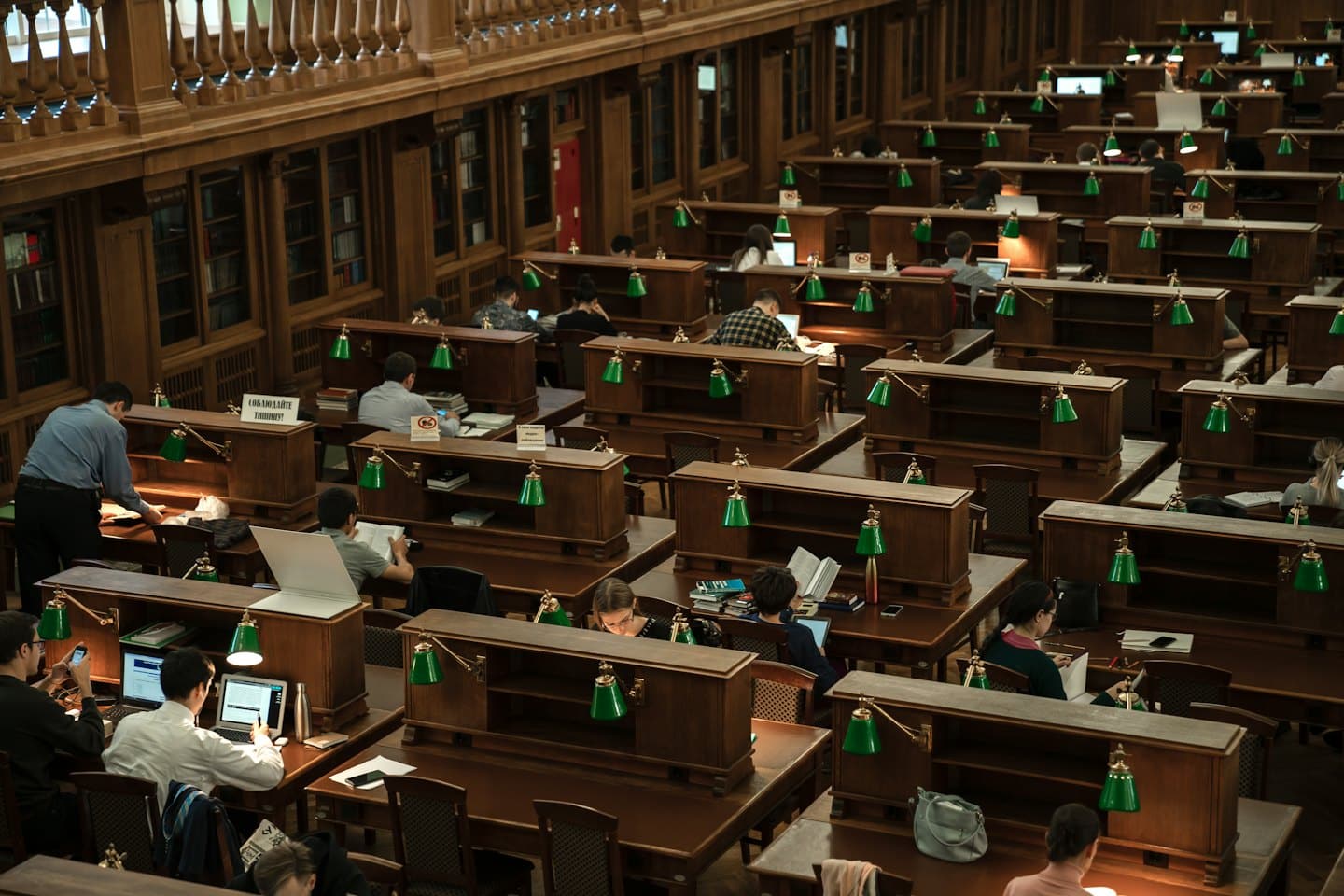 Students study at wooden desks with green lamps in a library.