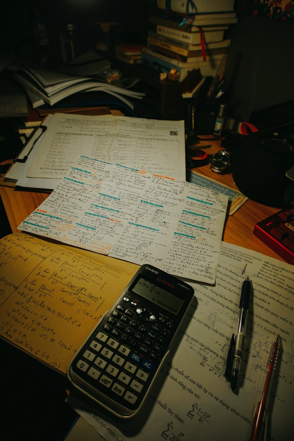 Notebooks, textbooks, and papers with math equations on a cluttered desk.